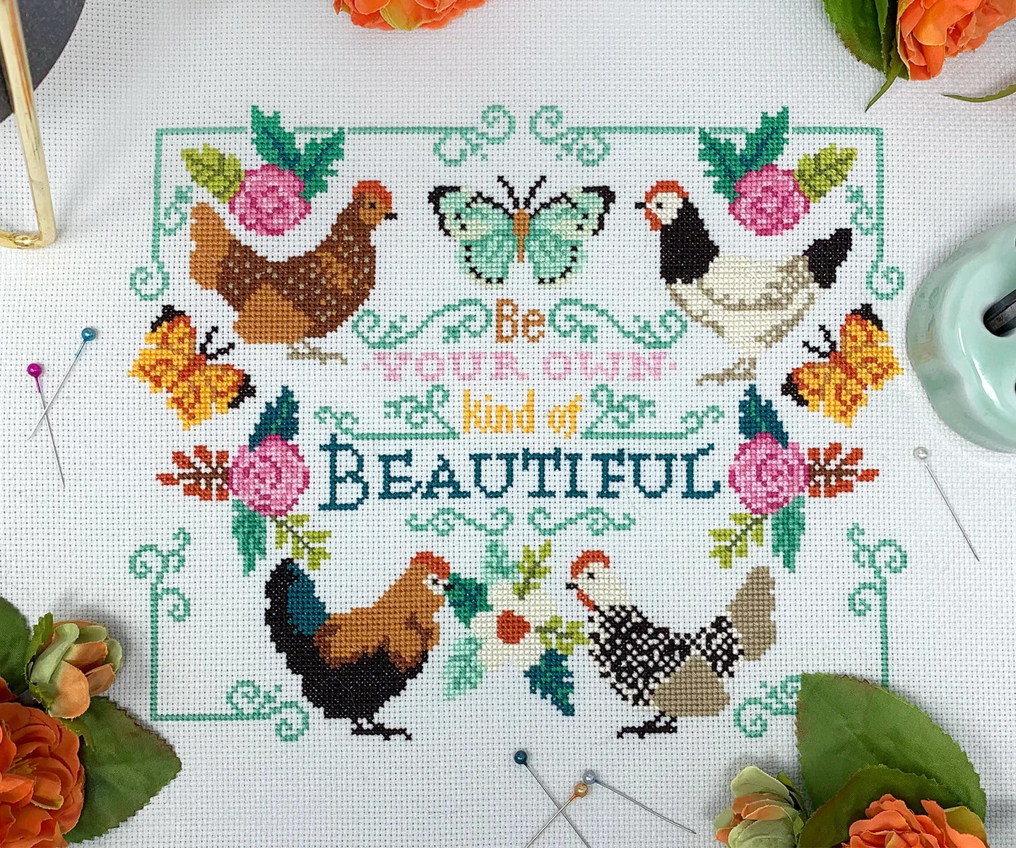 A cross stitch of colorful chickens, blooming flowers, fluttering butterflies, and the phrase “Be Your Own Kind of Beautiful” in the center.