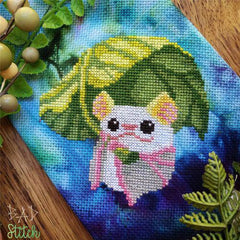 Cross stitch of a small bat holding a variegated green leaf over its head for shade, stitched on a bright blue tie-dyed background.