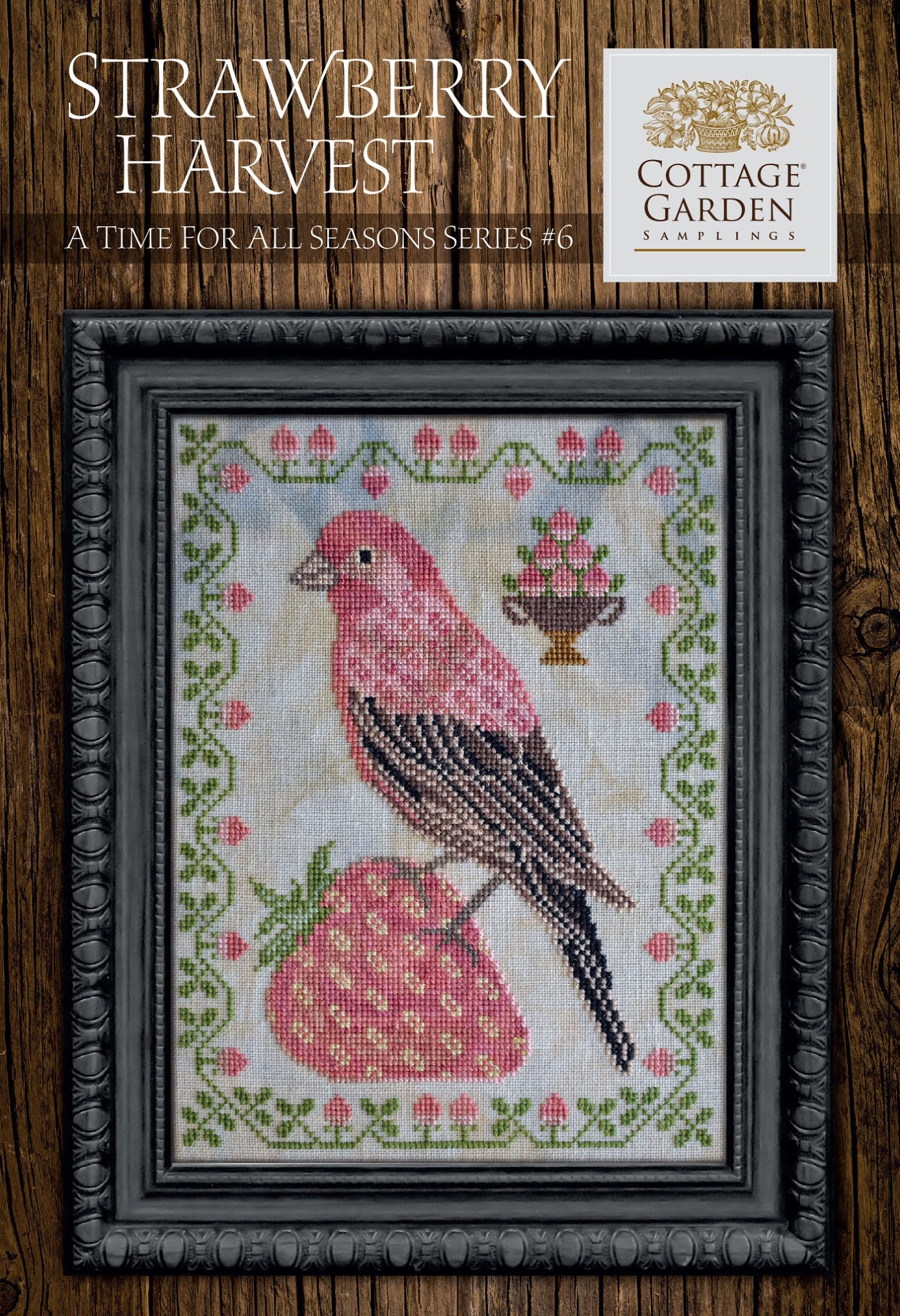 Framed cross-stitch sampler featuring a pink bird perched on a large strawberry, surrounded by a decorative border of green vines and small blossoms with a bowl of strawberries on a neutral background.
