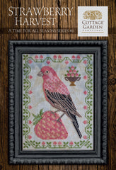 Framed cross-stitch sampler featuring a pink bird perched on a large strawberry, surrounded by a decorative border of green vines and small blossoms with a bowl of strawberries on a neutral background.
