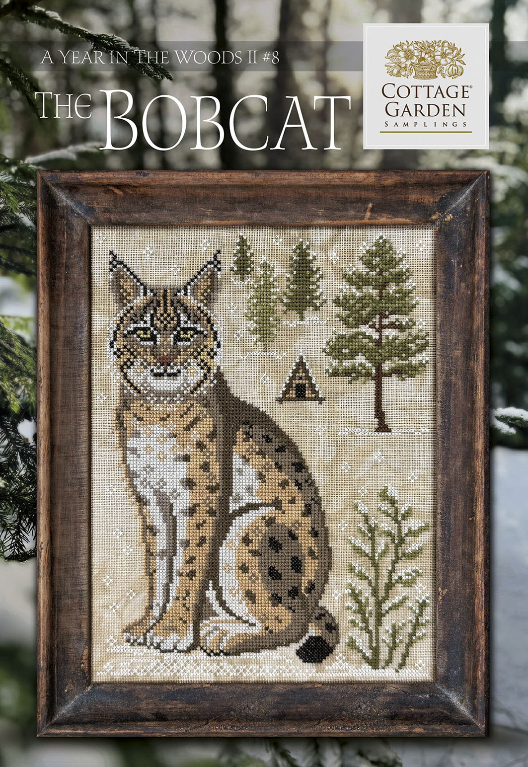 Framed cross stitch of a bobcat seated in a snowy woodland scene with evergreen trees and a small cabin.