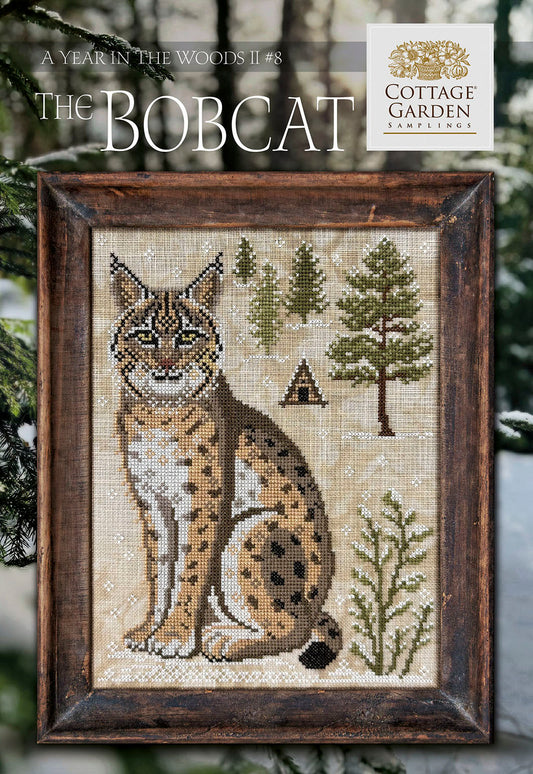 Framed cross stitch of a bobcat seated in a snowy woodland scene with evergreen trees and a small cabin.
