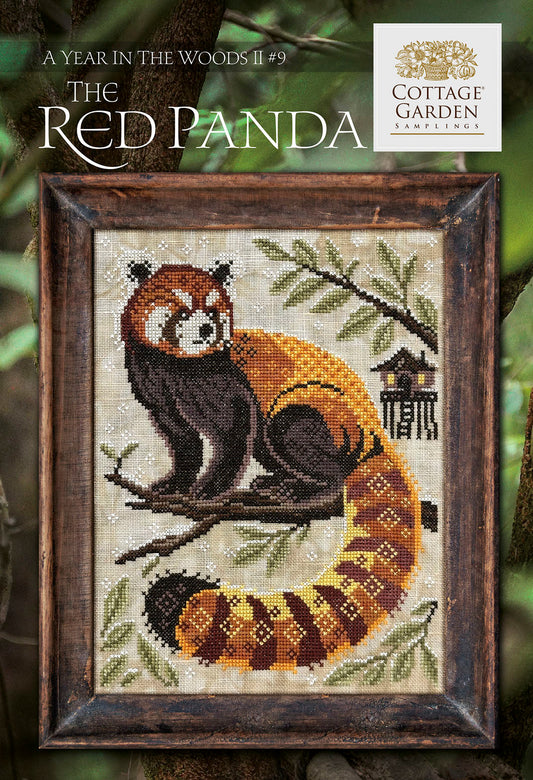 Framed cross stitch of a red panda curled on a tree branch in a forest scene, with pine branches and a small woodland structure nearby.