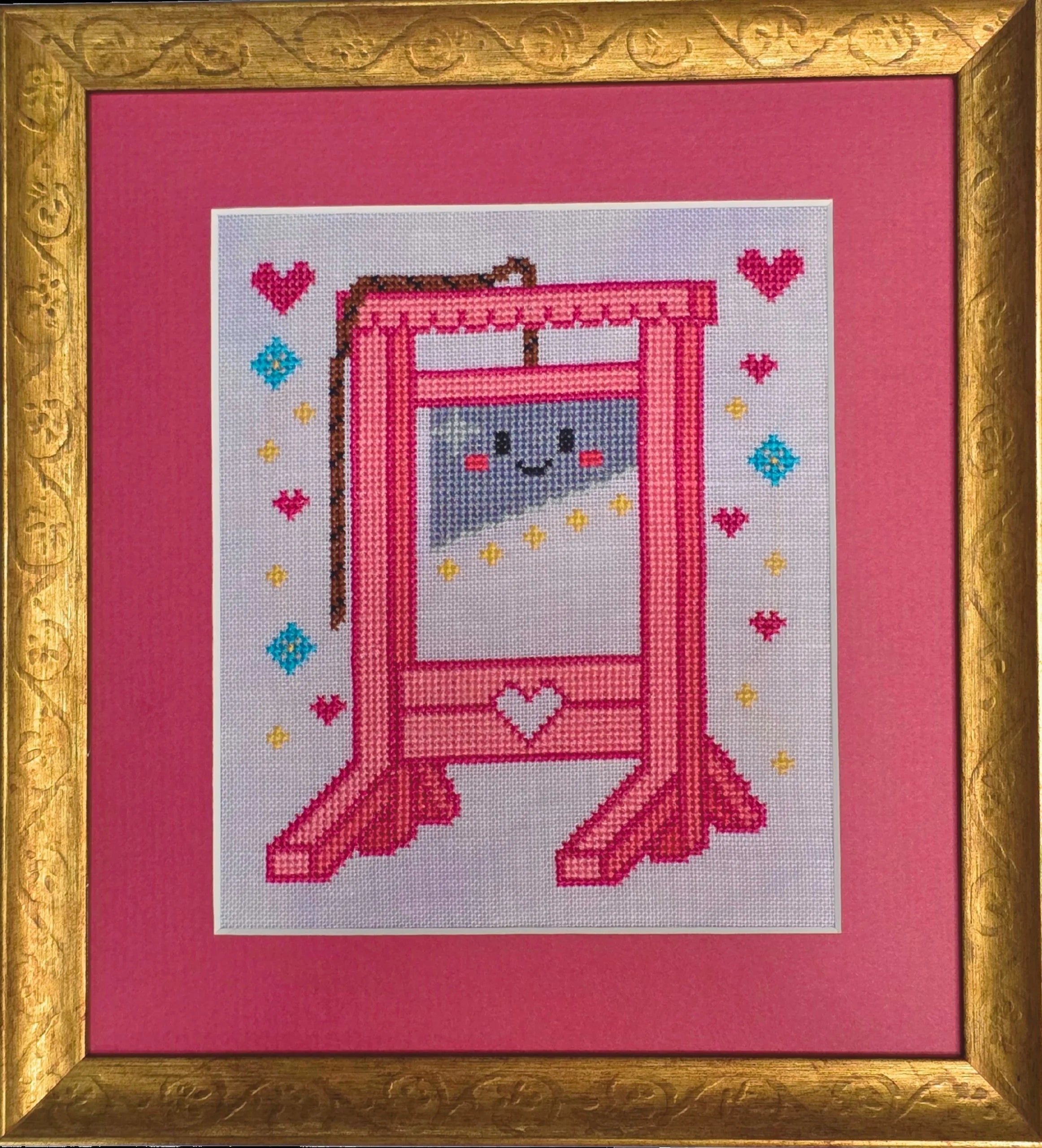 Framed cross stitch of a bright pink guillotine with a smiling cartoon face on the blade, surrounded by stitched hearts, stars, and diamonds on a light background, set within a gold frame and pink mat.