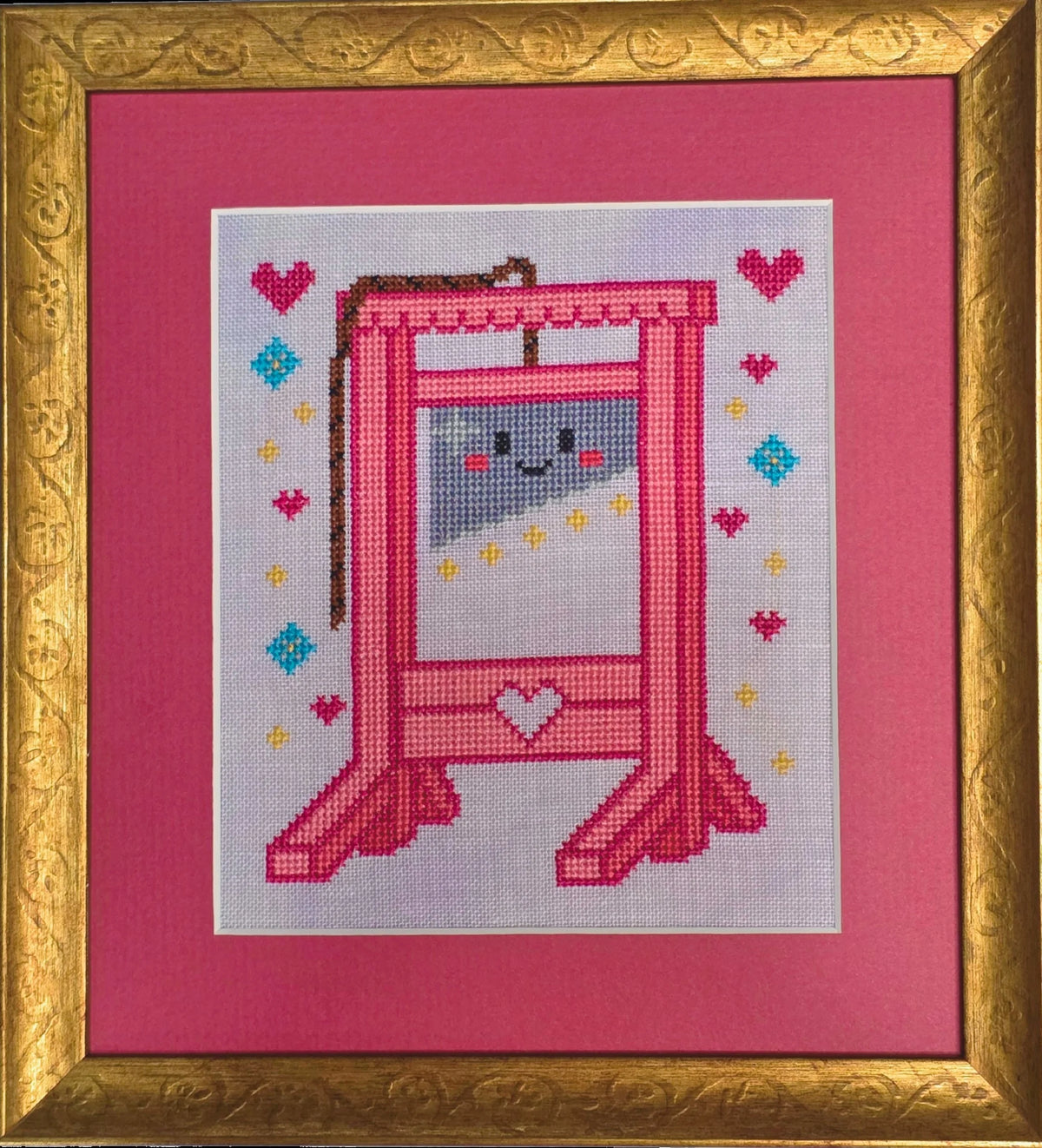 Framed cross stitch of a bright pink guillotine with a smiling cartoon face on the blade, surrounded by stitched hearts, stars, and diamonds on a light background, set within a gold frame and pink mat.