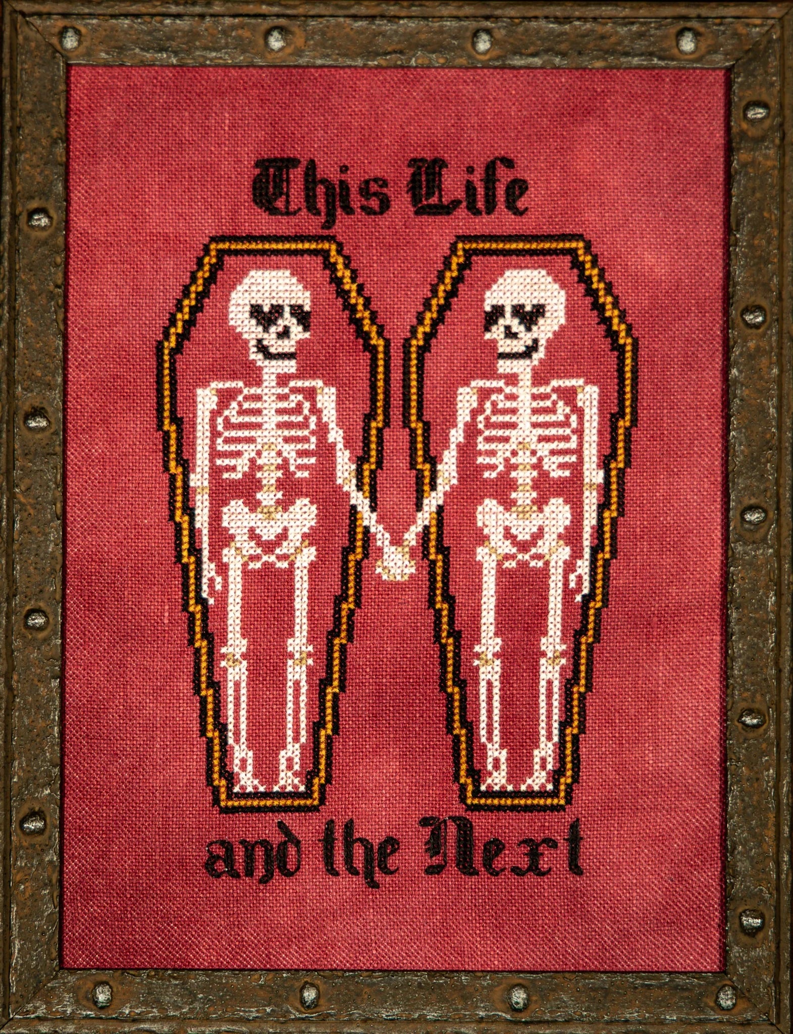 Cross stitch showing two skeletons lying in side‑by‑side coffins and holding hands, with the text “This Life” above and “and the Next” below, stitched on a red background and framed in a rustic metal‑style frame.
