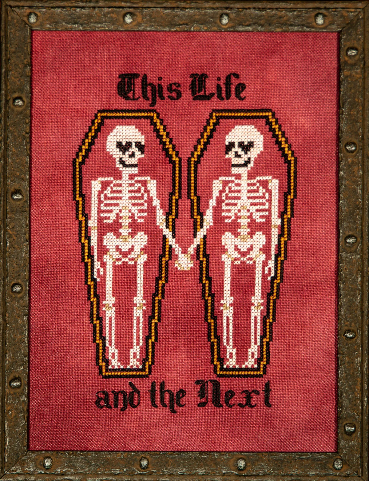 Cross stitch showing two skeletons lying in side‑by‑side coffins and holding hands, with the text “This Life” above and “and the Next” below, stitched on a red background and framed in a rustic metal‑style frame.
