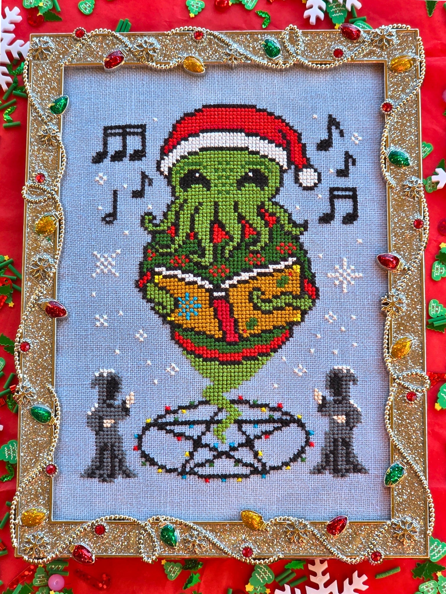 Cross stitch of a green tentacled Cthulhu creature wearing a Santa hat and singing from a book, surrounded by musical notes and snowflakes, with two robed figures below and framed in a gold tinsel‑like border decorated with colorful lights.