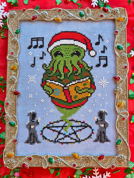 Cross stitch of a green tentacled Cthulhu creature wearing a Santa hat and singing from a book, surrounded by musical notes and snowflakes, with two robed figures below and framed in a gold tinsel‑like border decorated with colorful lights.