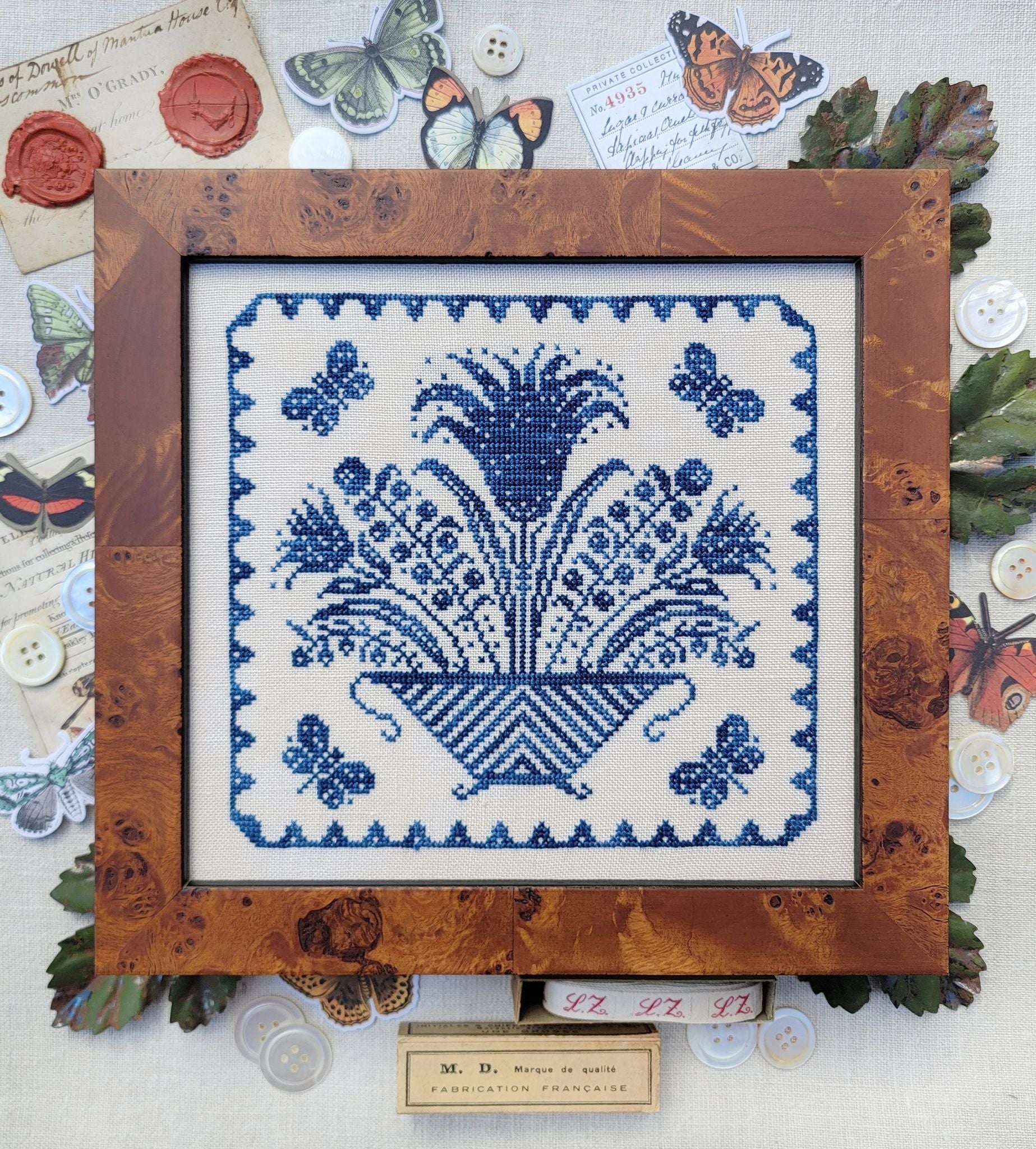 Framed cross stitch of a blue floral basket with butterflies, stitched in a traditional sampler style on light linen.