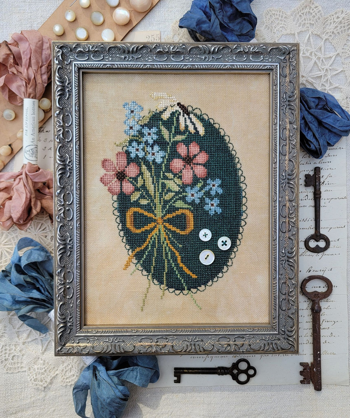 Framed cross stitch of a floral arrangement with pink and blue flowers tied with an orange ribbon with pearl button accents, with a green oval cross stitch background, set on natural linen.