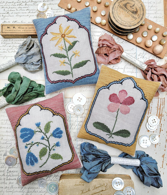 Three small cross stitched pillows featuring simple floral designs. A yellow flower, a pink flower, and blue blossoms - arranged with embroidery floss and vintage sewing notions.