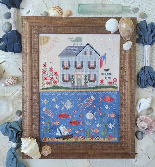 Framed cross stitch of a seaside house above an underwater scene filled with colorful fish, shells, and sea plants.
