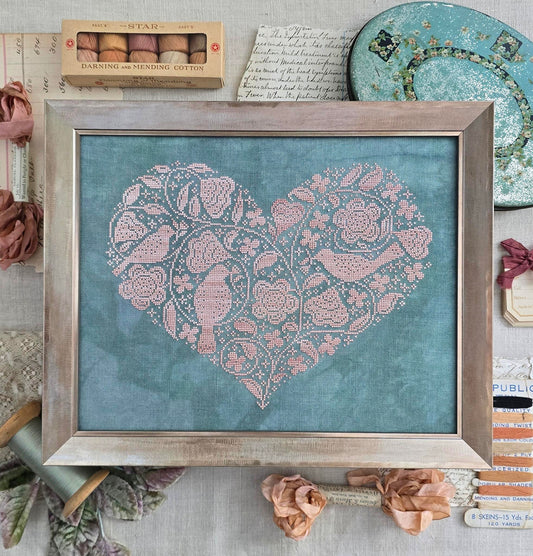 Framed cross stitch of a large pink heart filled with bird and floral motifs on a blue‑green fabric background.