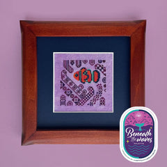 Framed cross stitch of an orange clownfish swimming among purple anemone‑like patterns on a lavender background.
