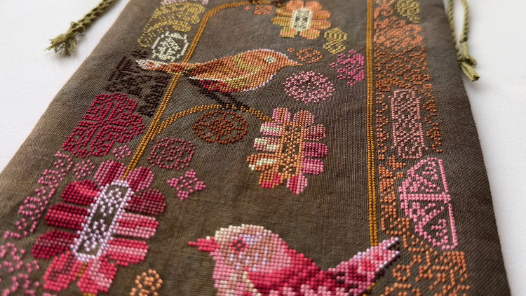 Close up of cross stitch wall hanging featuring stylized birds perched among pink and orange flowers on a dark fabric background.
