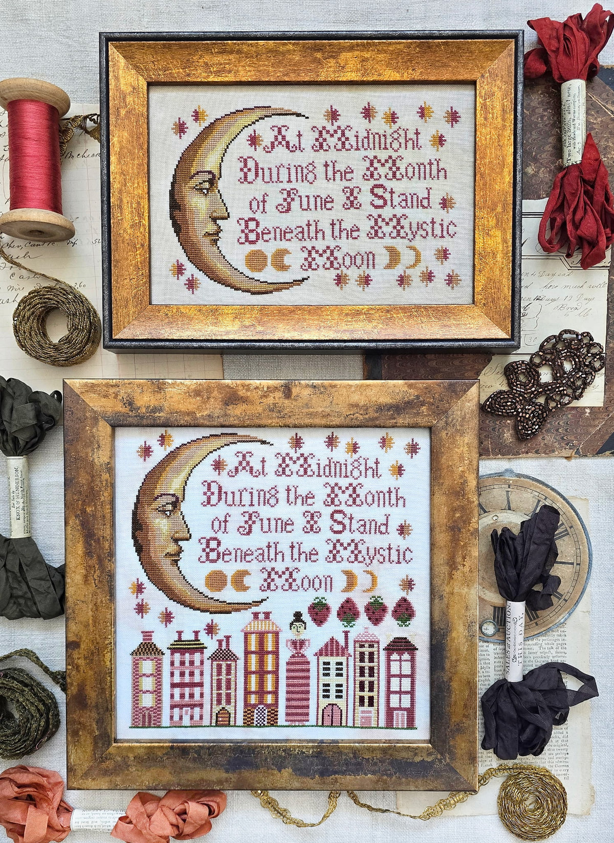 Framed cross-stitch samplers featuring a crescent moon face with decorative stars and stitched verse, with one design above a row of stylized houses, displayed with vintage sewing notions and ribbon.
