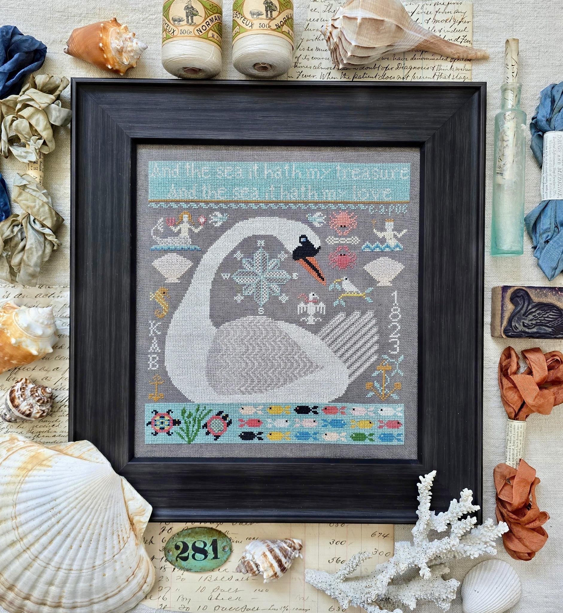 Framed cross-stitch sampler of a large white whale surrounded by small ocean motifs, including fish, shells, an anchor, a compass, and a bird, against a gray background with decorative borders.
