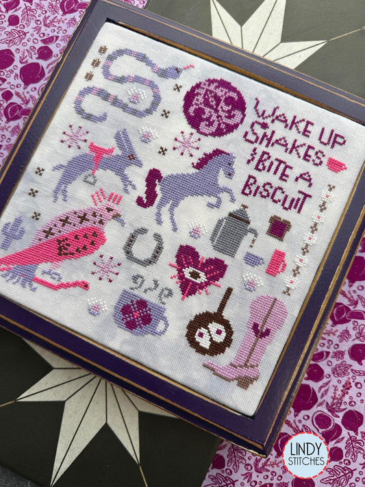Right tilted angle view of framed cross stitch in pinks and purples with a horses, a rabbit, a snake, a bird, boots, a coffee pot, and the phrase “Wake up snakes, bite a biscuit.”