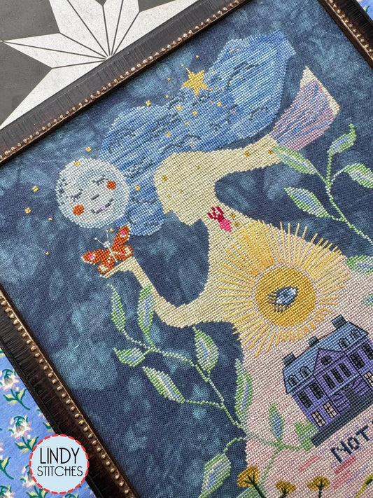 Close‑up of the upper portion of the cross‑stitch featuring the flowing blue hair, moon face, butterfly, and sunburst details.