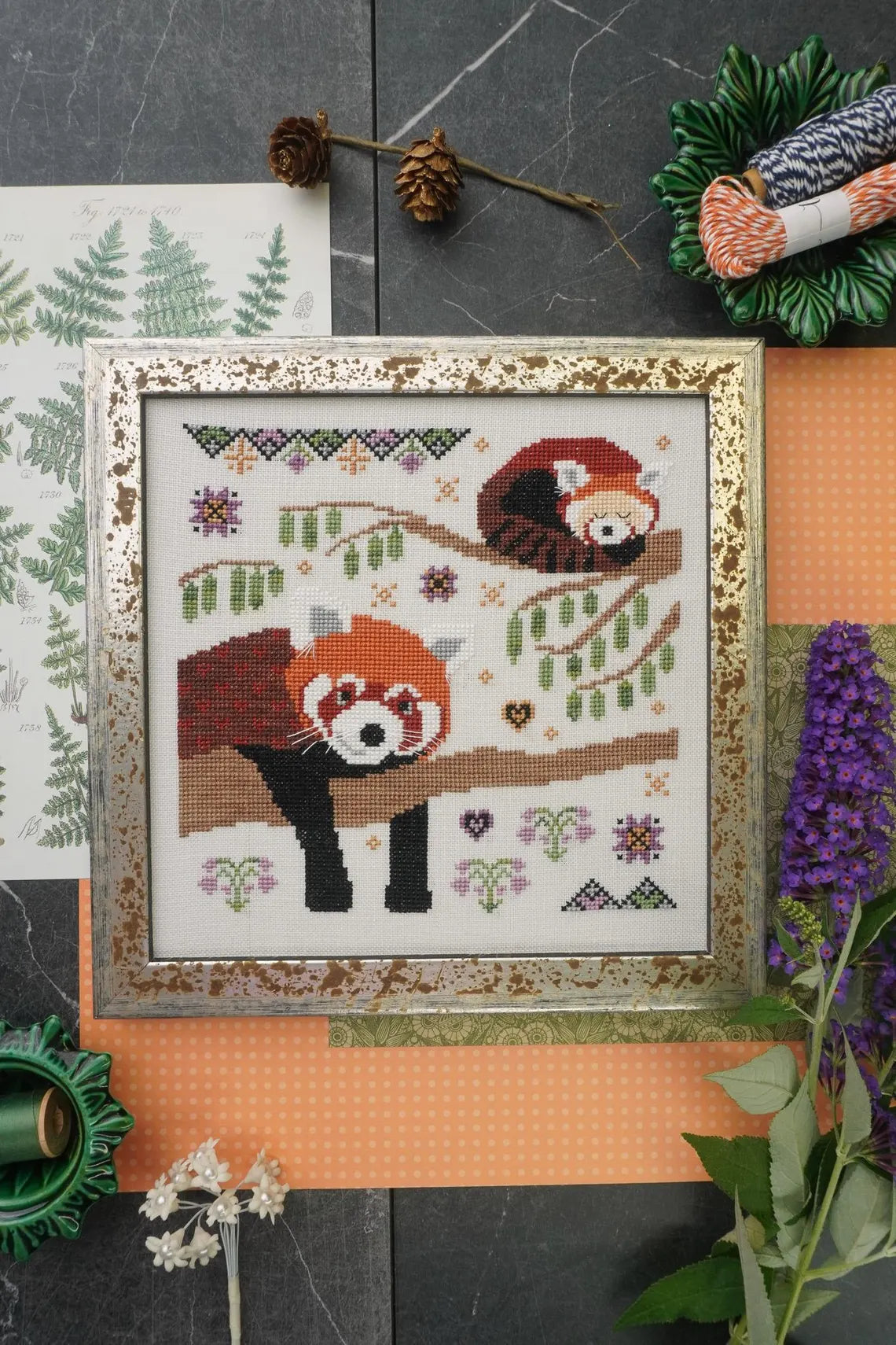 Framed cross‑stitch of two red pandas lounging on tree branches surrounded by small floral and geometric motifs.