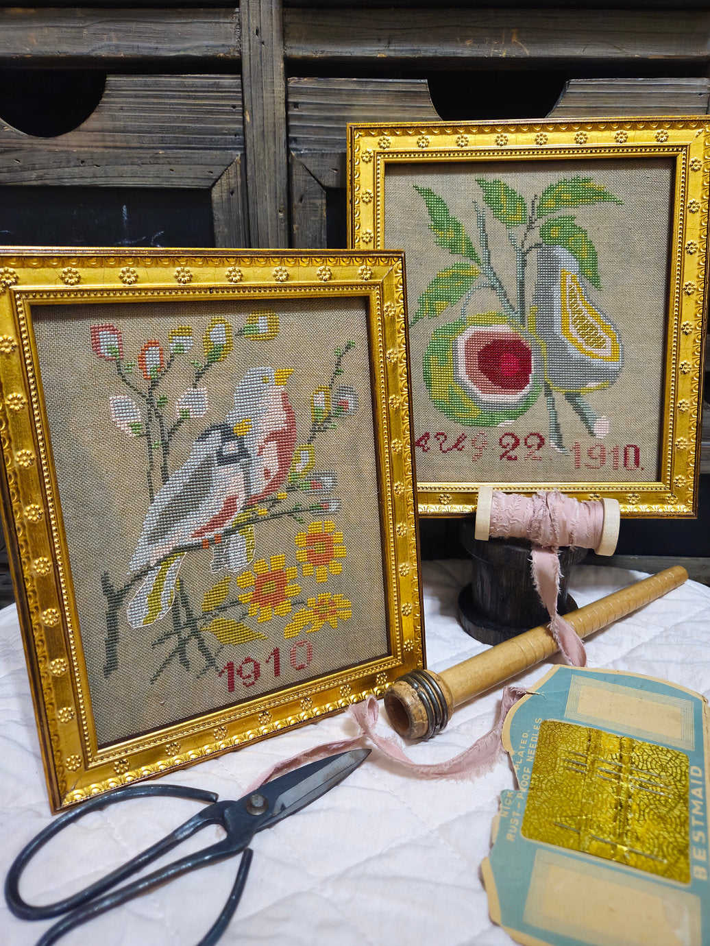 Two ornate gold-framed cross‑stitch pieces displayed on a wooden surface, one showing birds and flowers with ‘1910’ stitched below, the other depicting a pear and fig with ‘Aug 22 1910.’ Surrounding them are vintage sewing items including scissors, pink ribbon, a wooden bobbin, and a packet of rust‑proof needles.