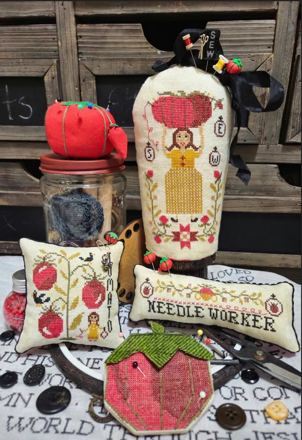 Collection of cross‑stitched sewing-themed pieces, including a figure holding a large tomato needle cushion, pincushions with tomato motifs, and small cross stitched pillows reading 'TOMATO' and 'NEEDLE WORKER' arranged with sewing tools and buttons.