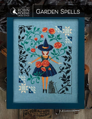 Framed cross-stitch design titled “Garden Spells” featuring a central figure in a pointed hat and dark dress surrounded by orange flowers, green vines, black botanical silhouettes, geometric quilt blocks, and a small gray cat on a blue background.
