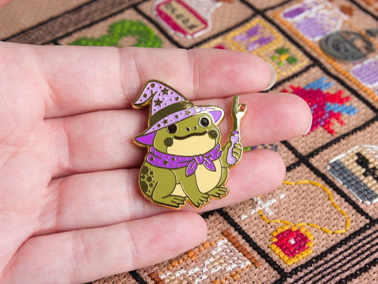 Enamel needle minder of a cute frog wizard in a purple hat and cape, shown held in a hand on a patterned backing card over a colorful cross stitched background.