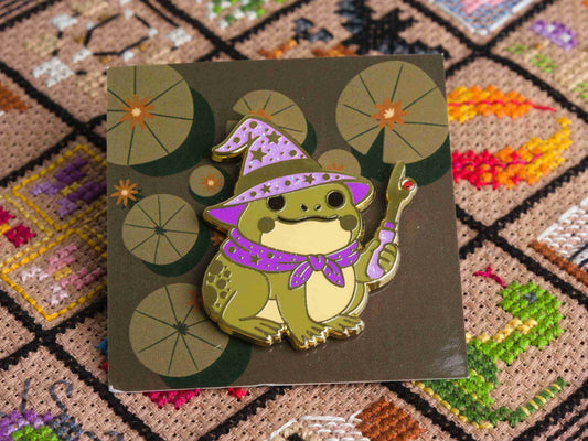 Enamel needle minder of a cute frog wizard in a purple hat and cape, shown on a patterned backing card over a colorful cross stitched background.