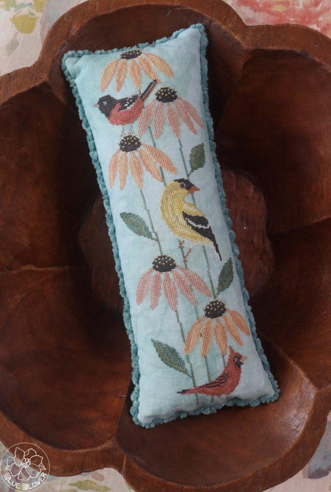 Long, narrow cross‑stitched pillow featuring three birds perched among tall coneflower blooms, displayed inside a carved wooden bowl.
