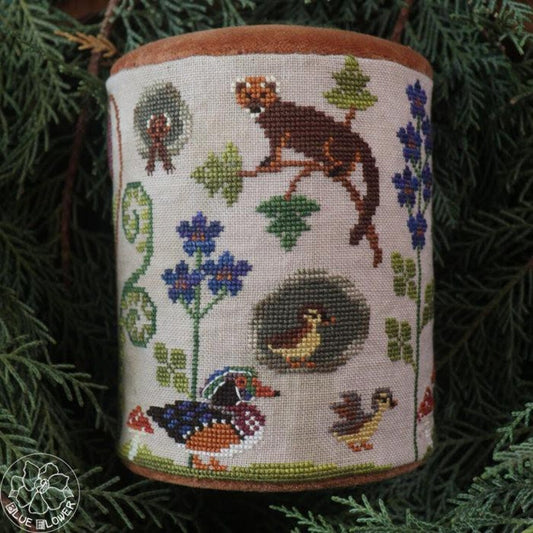 Cross stitched cylindrical drum featuring woodland animals, including a small animal perched on a branch, ducks, and chicks among green plants and blue flowers.