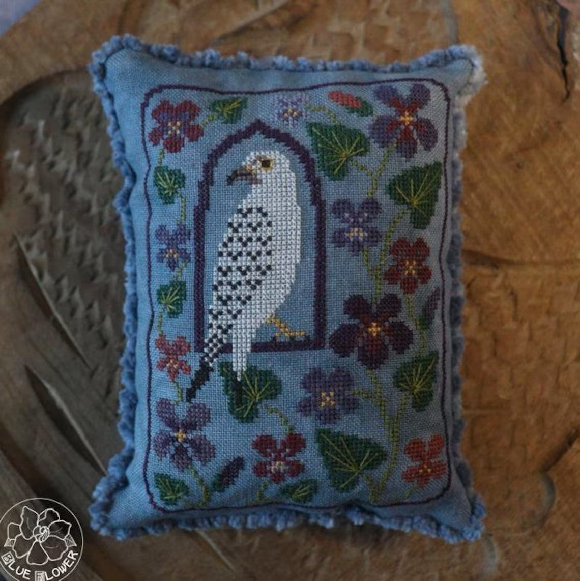 Cross stitched pillow featuring a white falcon framed in an arch, surrounded by purple and pink flowers on a blue fabric background.