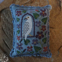 Cross stitched pillow featuring a white falcon framed in an arch, surrounded by purple and pink flowers on a blue fabric background.