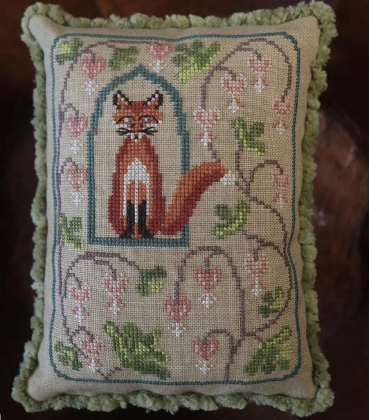 Cross stitch pillow featuring a red fox framed in an arch window, surrounded by curling vines and pink bleeding flower blossoms, with a green ruffled edge.