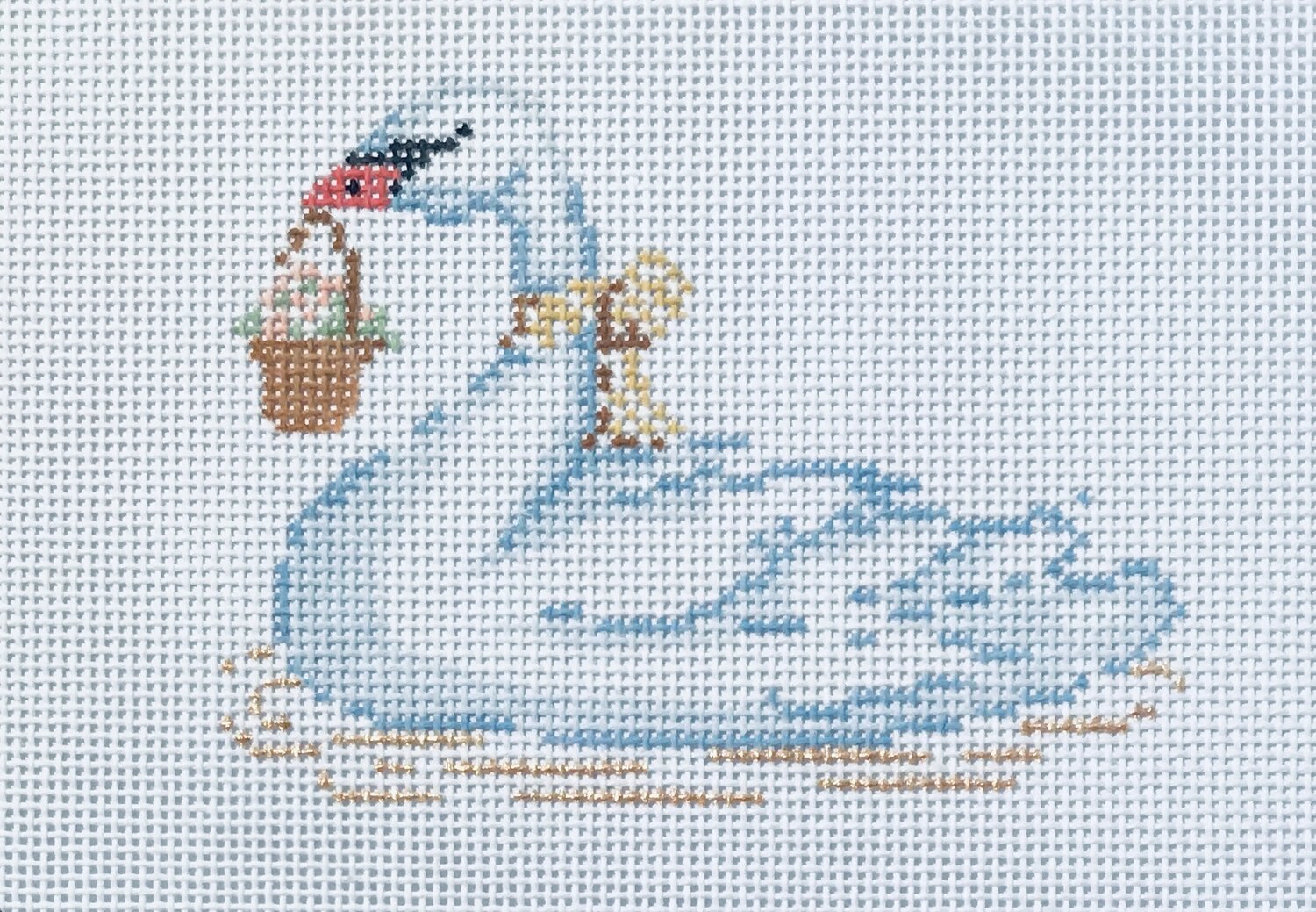 Needlepoint canvas featuring a white swan gliding across blue water, wearing a gold ribbon around its neck and carrying a basket of flowers.
