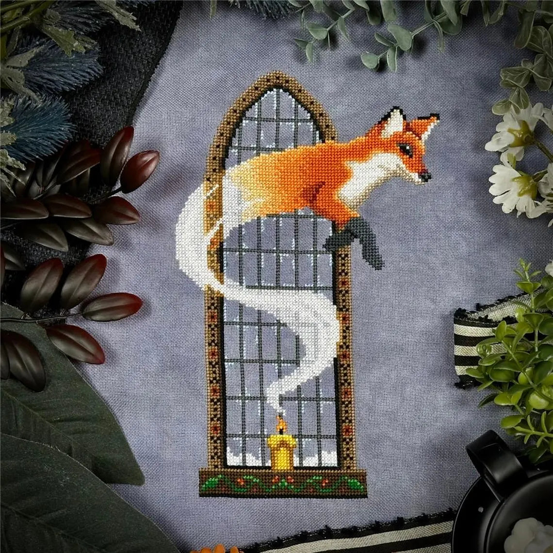 Cross‑stitch of a fox emerging in a swirl of white smoke from a lit candle in front of a tall arched window, surrounded by leafy props.
