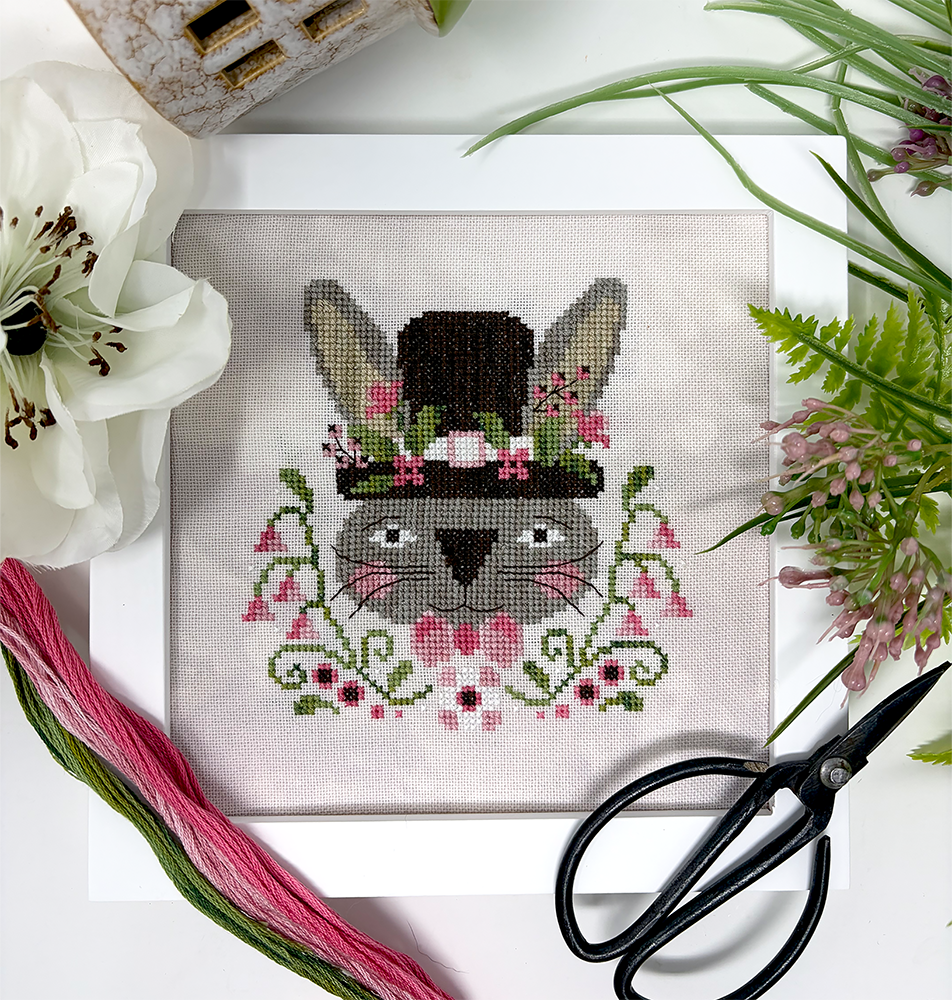 Cross-stitch design of a gray bunny wearing a tall top hat decorated with pink flowers, surrounded by green vines and blossoms, displayed on neutral fabric with embroidery floss, scissors, and flowers nearby.
