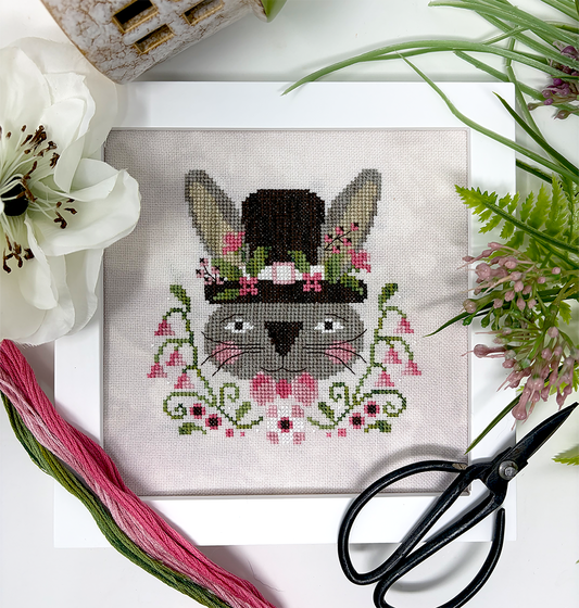 Cross-stitch design of a gray bunny wearing a tall top hat decorated with pink flowers, surrounded by green vines and blossoms, displayed on neutral fabric with embroidery floss, scissors, and flowers nearby.
