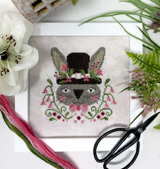 Cross-stitch design of a gray bunny wearing a tall top hat decorated with pink flowers, surrounded by green vines and blossoms, displayed on neutral fabric with embroidery floss, scissors, and flowers nearby.
