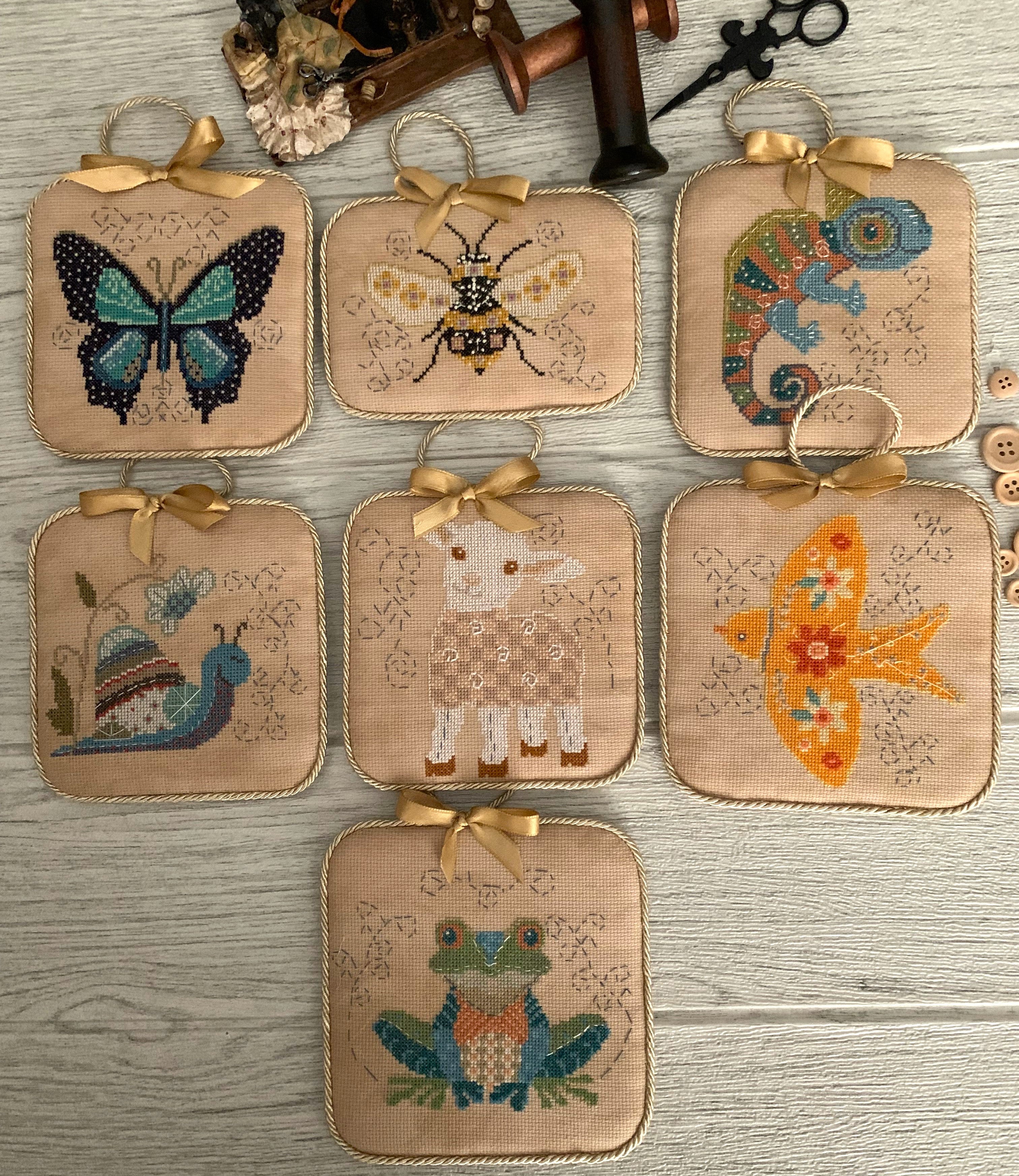 Set of seven small square cross stitch ornaments featuring animals and insects: a butterfly, bee, chameleon, snail, sheep, swallow, and frog. They're finished with ribbon hangers on natural fabric.